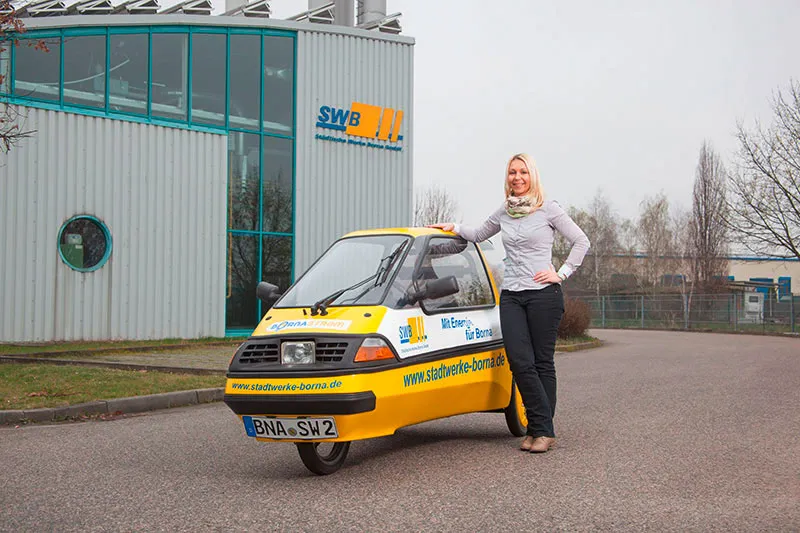 Eine Frau steht neben einem kleinen gelben dreirädrigen Auto mit Firmenlogo, das vor einem Industriegebäude geparkt ist.