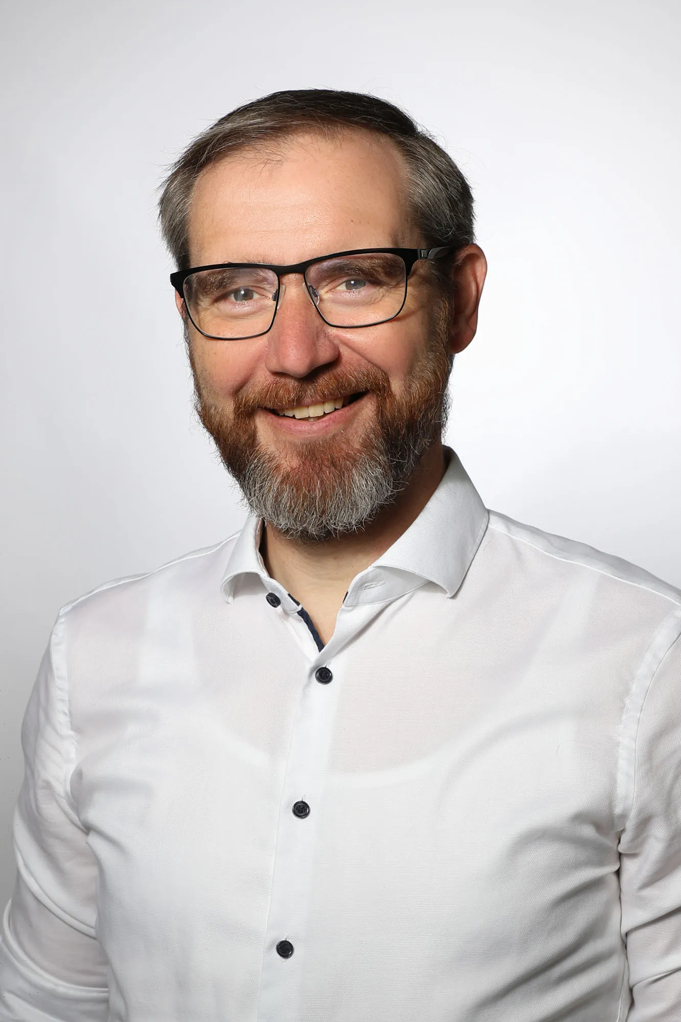 Ein Mann mit Brille und Bart lächelt in die Kamera. Er trägt ein weißes Button-up-Hemd vor einem schlichten hellen Hintergrund.
