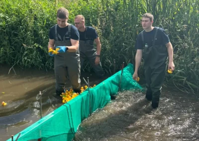 Drei Personen in Wathosen sammeln Gummi-Enten aus einem Netz in einem Bach, während andere Personen und ein Feuerwehrauto am grasbewachsenen Ufer im Hintergrund zu sehen sind.