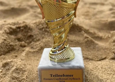 Eine goldene Trophäe mit der Aufschrift Teilnehmer Firmen Cup Bona City Beach 2024 steht auf sandigem Boden, während Menschen und Ausrüstung im Hintergrund verschwommen zu sehen sind.
