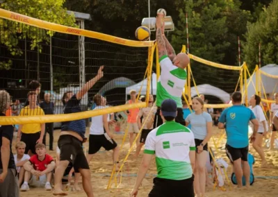 Ein Spieler springt, um den Ball über das Netz zu schlagen, während andere zusehen oder sich darauf vorbereiten, zu reagieren. Im Hintergrund sind Bäume und Zelte zu sehen.