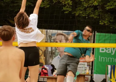 Zwei Teams spielen Beachvolleyball; ein Spieler im weißen Hemd springt, um den Spike eines Spielers im blauen Hemd zu blocken, während zwei andere zusehen.