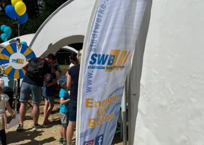 Eine Werbefahne der Stadtwerke Borna steht vor einem weißen Zelt bei einer Freiluftveranstaltung, im Hintergrund sind Menschen und Luftballons zu sehen.