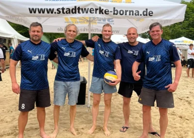 Fünf Männer in passenden blauen Sporttrikots stehen an einem Sandstrand unter einem Markenzelt und posieren für ein Gruppenfoto, wobei einer einen Volleyball hält.